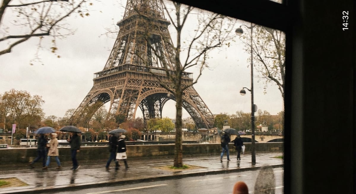 從移動的巴士車窗內俯瞰雨中的巴黎艾菲爾鐵塔，前景有數名撐著雨傘走在濕滑路面上的行人，整體色調呈現復古且憂鬱的氛圍。