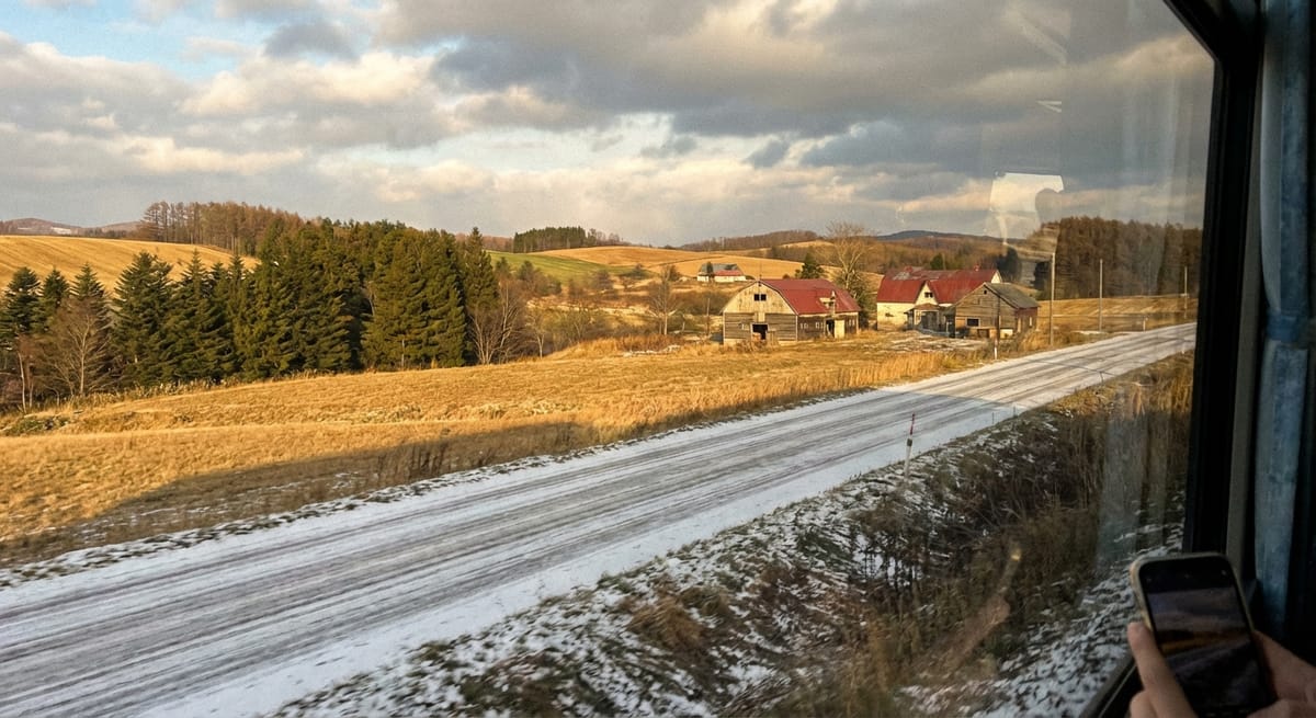 從移動的車窗向外拍攝北海道風景，畫面包含覆蓋薄雪的金色山丘、紅屋頂的木造農舍與松樹林，右下角可見一隻手拿著手機正在拍照。