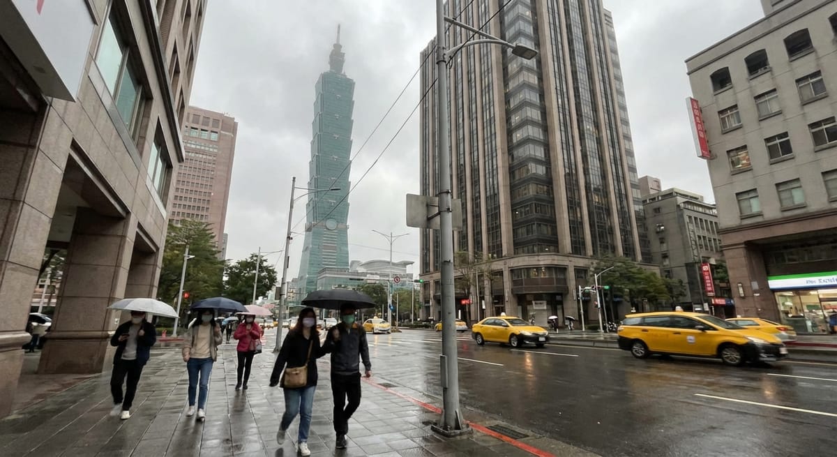 台北 101 在陰雨天的街景，行人撐著雨傘走在信義區濕漉漉的街道上，路面有黃色計程車行駛，兩旁為現代高樓建築。